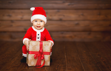 Happy baby in a Christmas costume Santa Claus with gifts on wooden backgroundの写真素材
