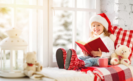 happy child girl reading a book while sitting at a winter window Christmasの写真素材