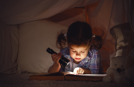 child girl reading  with  book and flashlight and teddy bear in tent. before going to bedの写真素材