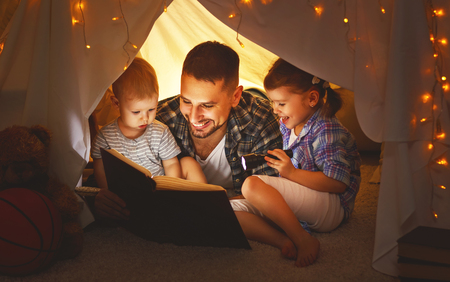 happy family father and children reading a book with a flashlight in a tent at homeの写真素材