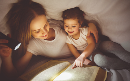 Mother and child daughter read book under a blanket with a flashlightの写真素材