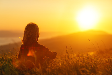 Woman sits with her back in the field and admires the sunset in the mountainsの写真素材