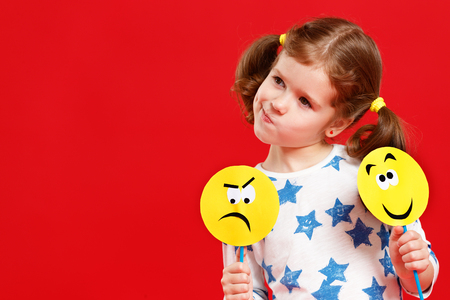 concept of children's emotions. child girl chooses between a sad and joyful smile on  colored red background
の写真素材