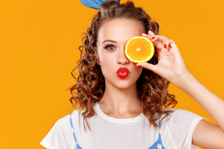 Cheerful young curly woman girl with orange on a yellow color backgroundの写真素材