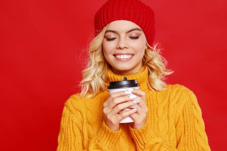 young happy emotional cheerful girl laughing  with knitted autumn cap and Cup of coffee  on colored red backgroundの写真素材