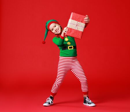 cheerful funny baby in Christmas elf costume with gifts on colorful red backgroundの写真素材