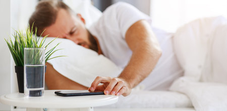 young bearded man wakes up in the morning in his bed with a phone alarm clock