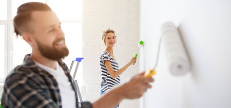 Cheerful couple  young woman smiling and watching boyfriend painting white wall with roller during renovation in new apartmentの写真素材