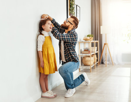 Side view of young man in casual clothes measuring height of little daughter next to white wall in light living roomの写真素材