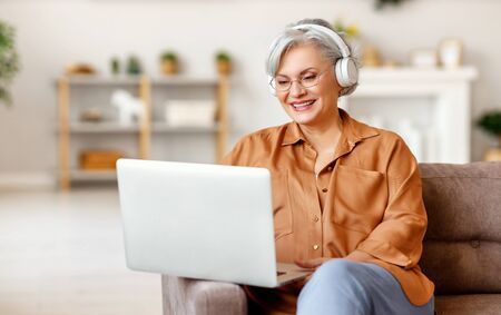 Happy senior lady in casual clothes and headphones smiling and watching movie on laptop while sitting on sofa at homeの写真素材