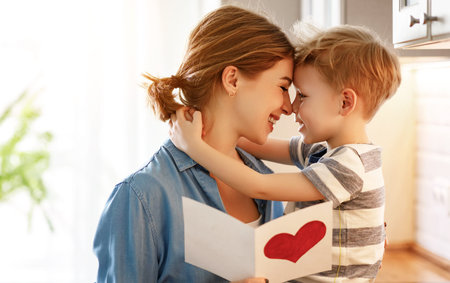 Happy little boy congratulating smiling mother and giving card with red heart during holiday celebration at homeの写真素材