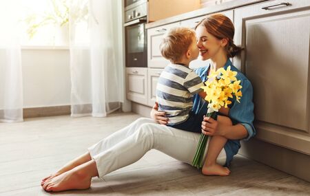Cheerful boy smiling and hugging happy woman with bouquet of flowers during holiday celebration sitting on the floor in kitchen at homeの写真素材