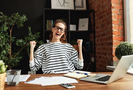 Happy young lady with eyes closed and arms raised rejoicing in success while sitting at wooden table with notepad and laptop in loft office among green potted houseplantsの写真素材
