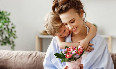 Happy young mother enjoying presented bouquet of delicate fresh flowers while caring small son hugging and congratulating with holiday at homeの写真素材