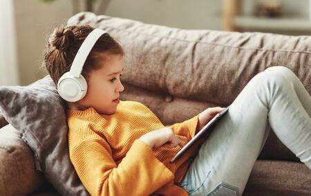 Full body happy girl in casual clothes and headphones s  playing game on tablet while sitting on soft couch at homeの写真素材