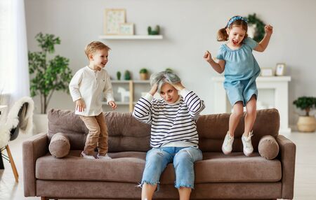 Shocked tired grandmother in casual wear sitting on sofa with hands on head while energetic grandchildren having fun and jumping around at homeの写真素材
