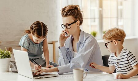 Bored small boy and pensive weary freelance mother in glasses focusing on screen of laptop while sitting at table with smartphone beside daughter drawing in notepad in cozy living room
の写真素材