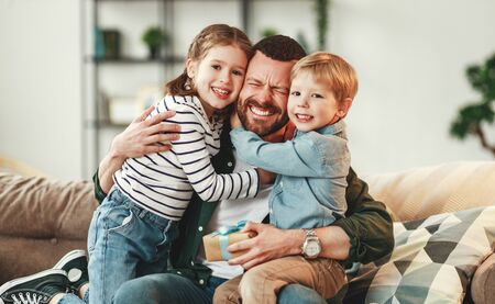 Adorable smiling little sister and brother hugging happy dad with gift box in hand while giving congratulations on fathers day at homeの写真素材