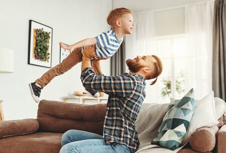 Cheerful father sitting on sofa and throwing up delighted little boy while having fun and enjoying time together in modern living roomの写真素材