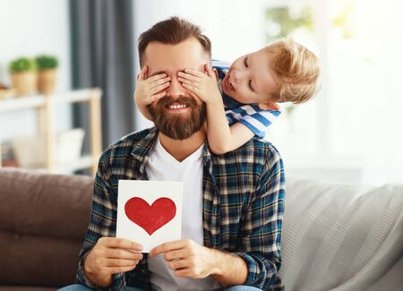 Happy young bearded man in casual wear with eyes closed and greeting card while sitting with little son on sofa at home in fathers dayの写真素材