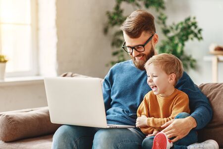 Cheerful bearded man in glasses hugging laughing boy and browsing laptop while sitting on sofa at home togetherの写真素材
