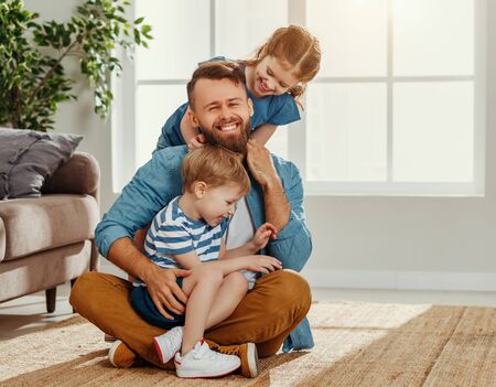Cheerful young man enjoying pastime with little son and daughter while sitting on floor and hugging in light living roomの写真素材