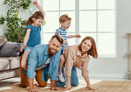 Cheerful young couple laughing   while having fun and giving piggyback ride to happy active kids in cozy living roomの写真素材
