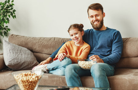 Smiling young dad with female kid sitting on couch against white wall in living room and cuddling while watching film during free timeの写真素材