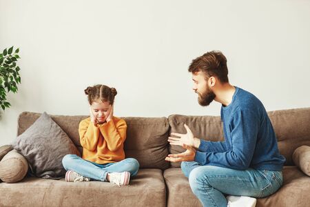 Bearded young man scolding daughter sitting on couch and gesturing while little girl sitting with legs crossed and eyes closed and covering earsの写真素材