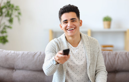 Happy young ethnic guy smiling and changing channels with remote control while sitting on couch and watching TV at homeの写真素材