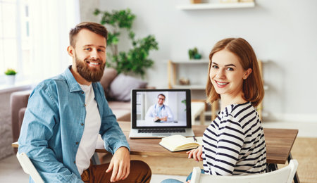 Cheerful young man and woman smiling and looking at camera while sitting at table at home and making...