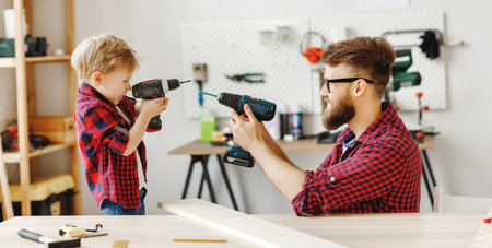Young bearded male and little boy in similar checkered shirts holding drills in modern carpentry...