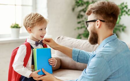 Young bearded man in glasses arranging bow tie on happy little son with backpack and books while preparing for school at homeの写真素材