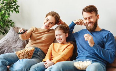 Optimistic family in casual wear sitting on sofa in light room and laughing while watching TV and...
