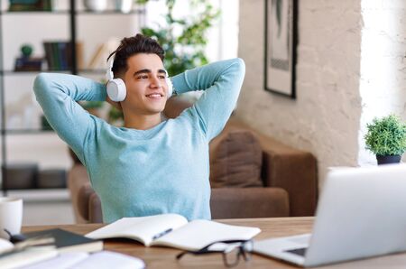 Positive young ethnic man keeping hands behind head and looking away while listening to music in headphones and resting after studies at homeの写真素材