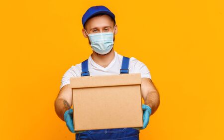 Male courier in protective mask and gloves giving carton box to camera while delivering order against yellow backgroundの写真素材