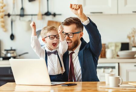 Young male entrepreneur in formal clothes and eyeglasses with little son in formal wear looking at laptop screen and celebrating successful deal while working remotely in modern apartmentの写真素材