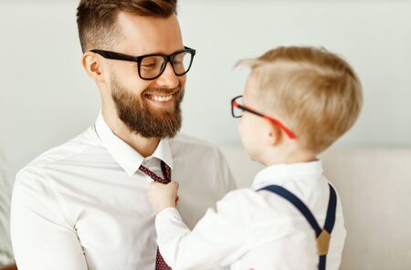 Back view of young child in formal wear and eyeglasses adjusting necktie of cheerful father in similar clothes and glasses while preparing together for event
の写真素材