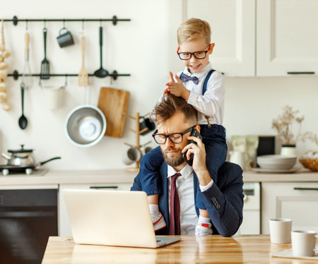 Cheerful playful kid sitting on neck of unhappy busy dad in formal wear during phone conversation and working with laptop in home kitchenの写真素材