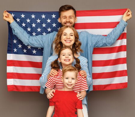Happy parents with USA flag and cheerful children hugging each other and looking at camera while celebrating Independence Day against gray backgroundの写真素材