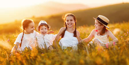 Ð¡heerful little kids in casual white clothes sit in the grass, having fun and laugh merrily in together in field at sunset while enjoying summer holidays in countrysideの写真素材
