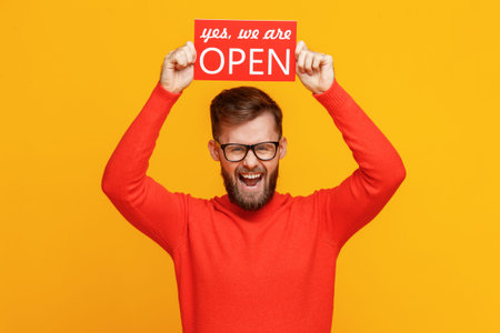 Happy bearded man smiling for camera and showing signboard with Open inscription against yellow...