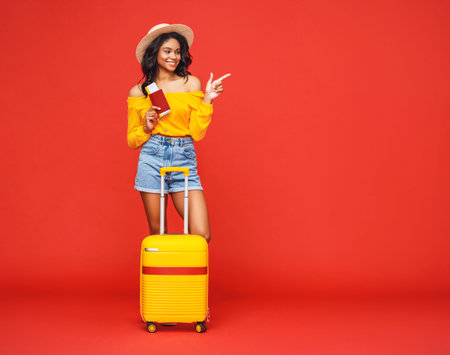Full body ethnic woman with tickets and suitcase smiling and pointing away during summer trip...