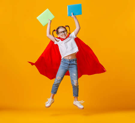 Full body happy schoolgirl in superhero cape smiling and holding notepads in raised arms during studies against yellow backgroundの写真素材