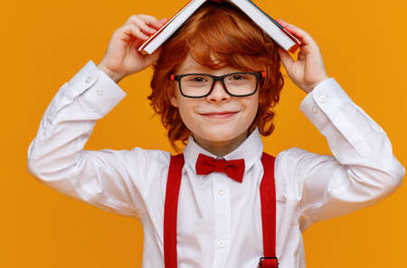 Delighted ginger boy in glasses smiling for camera and holds books on his head while having fun during school studies against yellow backgroundの写真素材