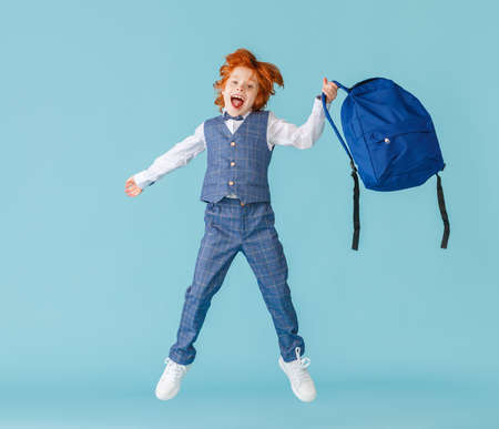 Full body delighted boy in school uniform screaming and leaping up with backpack against blue backdropの写真素材