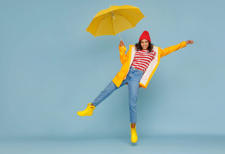 Full body happy ethnic woman in stylish outfit carrying umbrella and jumping on blue backgroundの写真素材