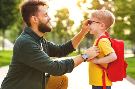 Supportive man adjusts glasses to cheerful smart schoolboy while preparing son to school in green summer parkの写真素材