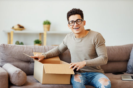 Happy ethnic man in glasses smiling and unpacking delivery carton box with delivered goods while...