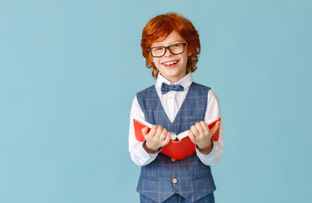 Happy redhead schoolboy in glasses smiling and looking at camera while reading book during school...
